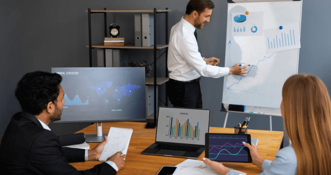 Man explaining a graph on a whiteboard to two colleagues, with charts and graphs displayed on nearby LCD screens during a target setting session.
